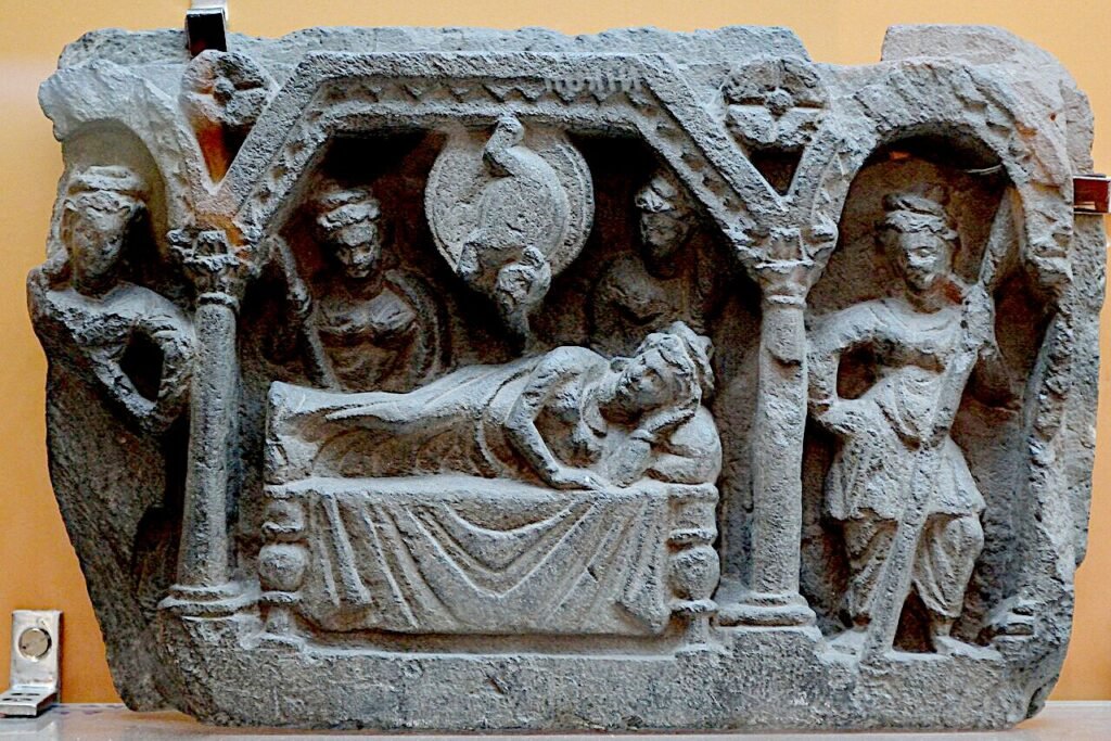 Gandhāran stone relief showing Queen Māyā reclining on a couch while a white elephant descends from above, symbolizing the Bodhisattva’s conception in early Buddhist narrative art.